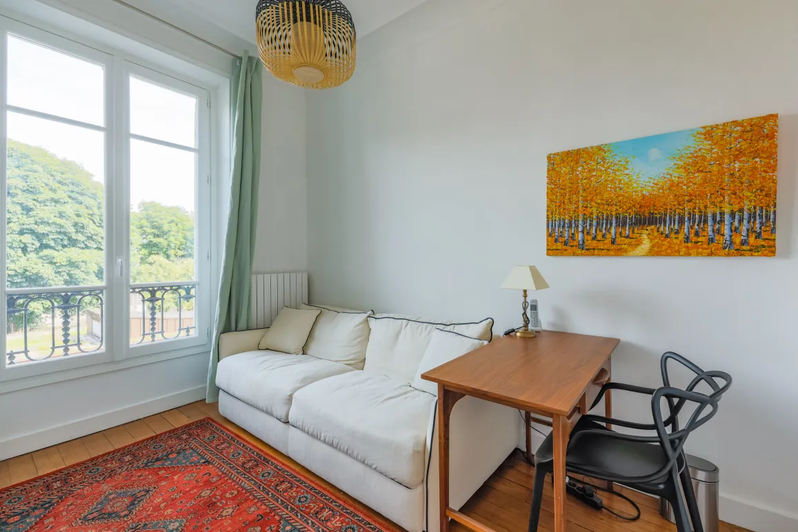 Bright room with green view, white sofa, wooden desk, and modern art. Serene, elegant ambiance for work or relaxation.