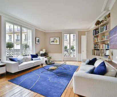 Bright luxury Parisian living room. White sofas, modern blue rug, hardwood floor. City view. Haussmannian elegance and comfort.