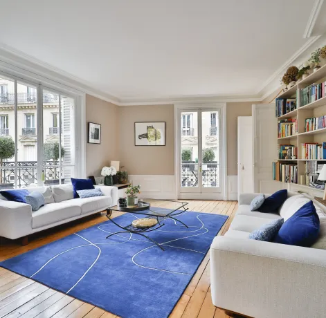Bright luxury Parisian living room. White sofas, modern blue rug, hardwood floor. City view. Haussmannian elegance and comfort.