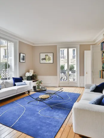 Bright luxury Parisian living room. White sofas, modern blue rug, hardwood floor. City view. Haussmannian elegance and comfort.